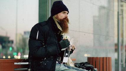 Homeless young man eating sandwich on bench at city street in evening