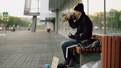 Homeless young man eating sandwich and drinking alcohol from paper bag on bench at city street in evening