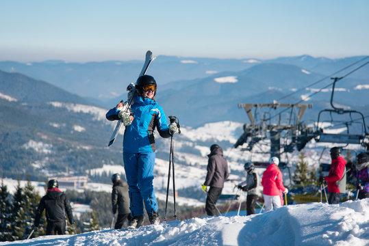 Full length shot of a happy woman posing with her skis on the shoulder on top of the hill, smiling joyfully. Blue sky, mountains, forests, ski lift and people on the blurred background