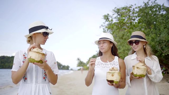several friends in hats walking along the beach and drinking juice from fresh coconut