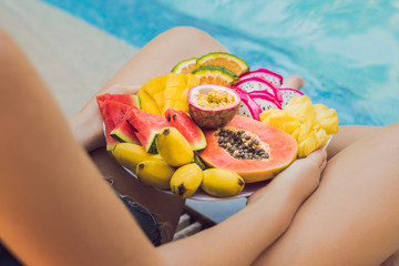 Young woman relaxing and eating fruit plate by the hotel pool. Exotic summer diet. Photo of legs with healthy food by the poolside, top view from above. Tropical beach lifestyle