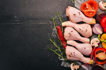 Raw chicken legs with fresh vegetables and rosemary and spices on a black wooden background. Top...