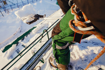 Team of male workers clean roof of building from snow with shovels in securing belts of mantra.