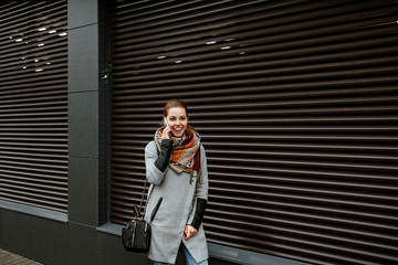 Street style. The girl communicates on the smartphone. The woman is dressed in jeans and a gray jacket, she costs on the street in the city against the background of an impressive gray wall.