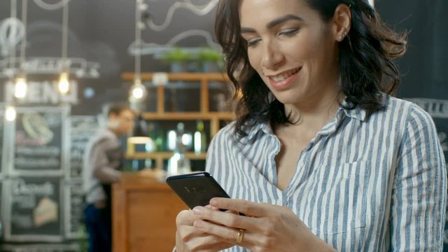 Beautiful Woman Sits in the Coffee Shop Uses Smartphone. She Smiles while Browsing Her Mobile Phone. In the Background Busy Stylish Cafe. Shot on RED EPIC-W 8K Helium Cinema Camera.