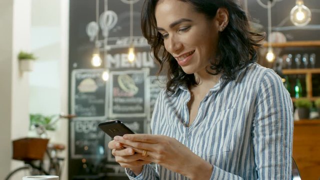 Beautiful Woman Sits in the Cafe Uses Smartphone. She Smiles while Messaging Her Friends or Loved One. On Her Table Cup with Beverage and Croissant. In the Background Busy Stylish Coffee House. 