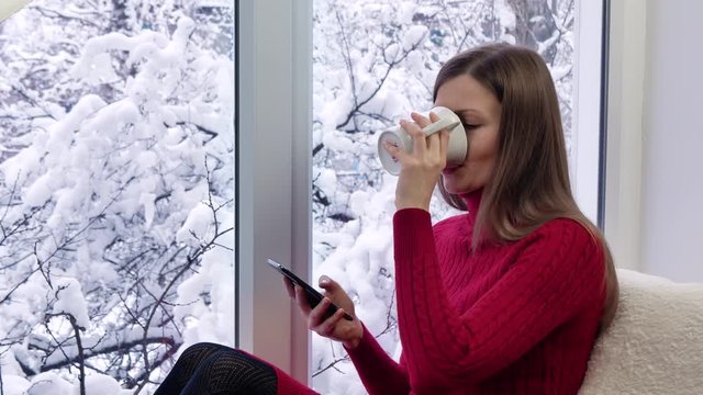 Pretty girl in red dress sitting on the windowsill, drinking tea and using smartphone. Winter outside.