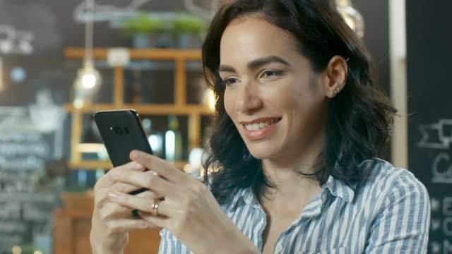 Portrait Shot of a Beautiful Young Woman Smiling and Using Mobile Phone. In the Background Stylish Cafe. Shot on RED EPIC-W 8K Helium Cinema Camera.