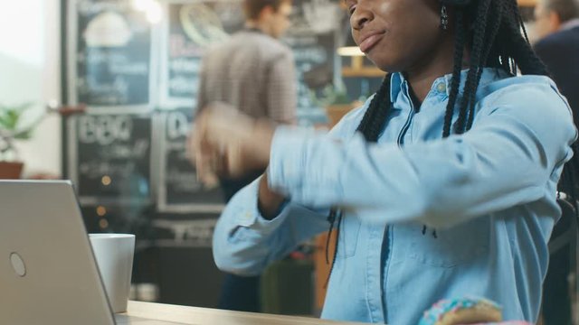 Stylish Young African-American Woman Sits in the Coffee Shop with Her Laptop and Headphones on, Listening to Music and Dancing to the Beats. Shot on RED EPIC-W 8K Helium Cinema Camera.