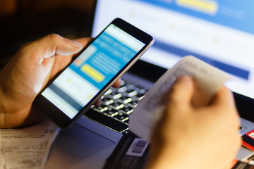 The smartphone app from a Bank to report cash and the tape is in the hands of the men in the background laptop. Man considers expenditures on a cash receipt.