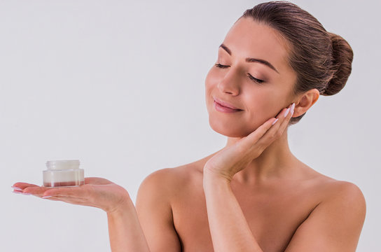 Confident in her beauty. Portrait of beautiful shirtless woman holding in one hand cream jar and touching face with another hand, isolated on white background. Skin treatment and beauty concepts