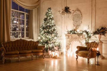Fototapeta na wymiar Christmas evening by candlelight. classic apartments with a white fireplace, decorated tree, sofa, large windows and chandelier.