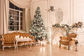 Christmas evening by candlelight. classic apartments with a white fireplace, decorated tree, sofa, large windows and chandelier.