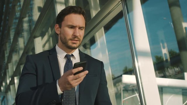 Caucasian handsome businessman in a suit and tie reading neaws on his black smartphone and getting very nervous, dissapointed and angry. Modern glass building. Indoors