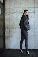 Obraz premium Beautiful lady with dark hair standing from back in classic gray suit and black shoes with heels and dreamily looking in camera 