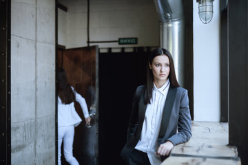 Portrait of beautiful girl with dark hair standing in classic gray suit and thoughtfully looking aside with girl that opening door on background