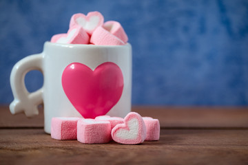 Close-up heart shape of pink marshmallow in white cup on wooden table with blue background. Concept for valentines day love celebration