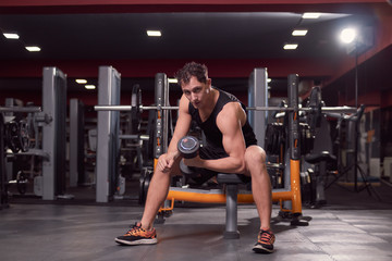 Fototapeta na wymiar one young man, dark gym indoors fitness equipment, one hand dumbbell biceps exercise, sitting bench, looking at camera.