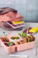 Gordijnen Lunch box with sandwiches, fresh fruits and vegetables on the table near school backpack © lithiumphoto