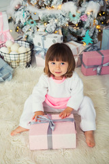 Funny baby girl in white and pink fleece pajamas sitting near Christmas tree and unpacking New Year gift