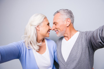 Close up photo of two happy cheerful lovely tender gentle sweet senior couple, they are taking a self portrait, hugging each other and touching noses, isolated on grey background