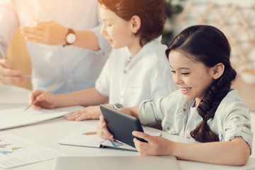 Modern generation. Cute joyful positive girl holding a tablet and smiling while sitting at the table