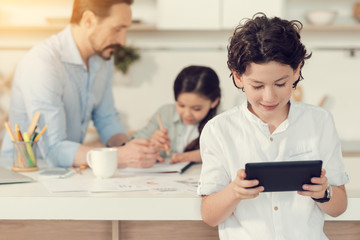 Obraz premium Modern igen. Nice positive smart boy smiling and looking at the tablet screen while using it for plying games