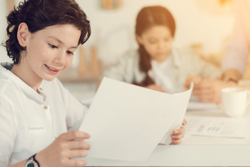 Smart children. Cute pleasant intelligent boy holding a piece of paper and reading his task while doing homework