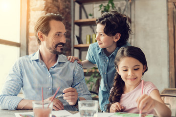 Time with family. Delighted positive handsome man sitting at the table and looking at his son while painting with him