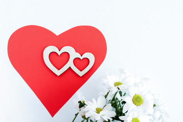 Red paper heart, two carved wooden hearts on it and white chrysanthemums on white background.