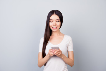 Portrait of attractive, young girl in white t-shirt holding telephone in her arms, looking at screen, using 5g internet, standing over grey background