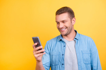 Bearded, cheerful, attractive guy in casual outfit, jeans shirt, holding smart phone in hand, using 5G internet, wi-fi, texting, chatting, writing sms, message over yellow background