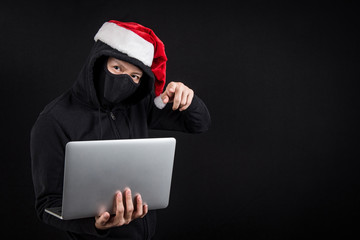 Mysterious male santa hacker holding laptop computer, anonymous man on black background, ransomware...