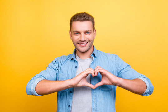 Portrait of  smiling, stunning man with stubble showing heart figure with fingers over yellow background
