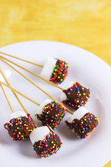 marshmallows on a sticks, laying on a white plate. glazed with chocolate and colorful sprinkles.