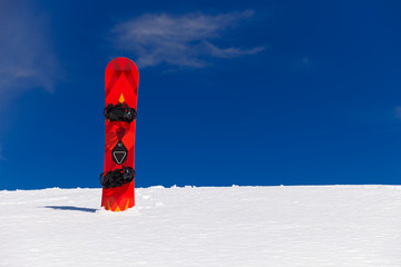 Orange snowboard with black duck stance positioned bindings plunged straight into snow with background of vivid blue sky
