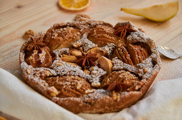 Just backed powdered pear pie decorated with white cloth, dried orange, walnuts, anise stars and spoon on the wooden board. Healthy homemade pear pie on the wooden background. Side view