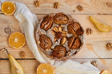 A powdered pear pie decorated with white cloth, fresh sliced pears, dried oranges, walnuts, anise stars, silver spoon on the wooden board. Healthy homemade pear pie. 