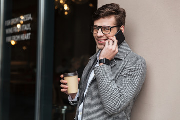 Cheerful business man in eyeglasses and coat talking by smartphone