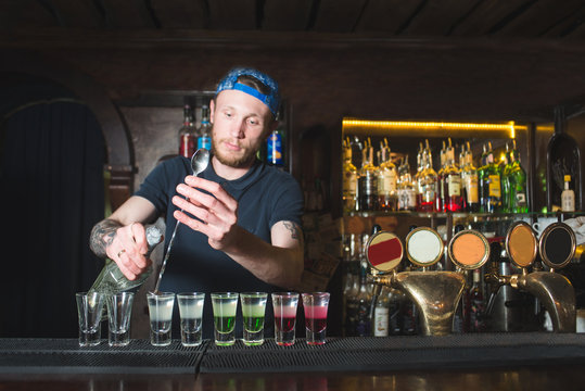 A barman with a beard makes colored alcoholic shots at the bar. Creation of showrooms at the bar restaurant.