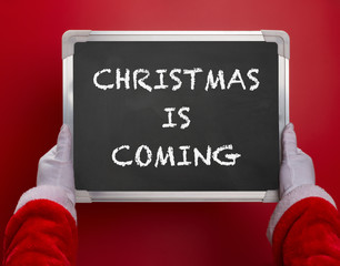 Fototapeta na wymiar first person angle view of Santa Claus holding a black chalk board written with CHRISTMAS IS COMING on red background