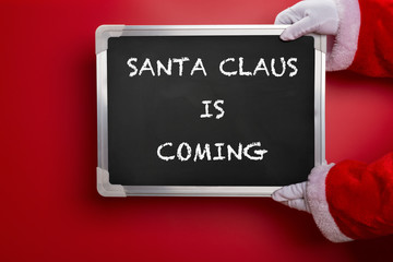 Santa Claus holding a black chalk board written with SANTA CLAUS IS COMING on red background