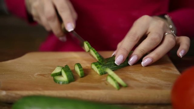 Woman's hands with perfect manicure slicing fresh cucumber on wooden cutting board in the kitchen. Closeup female hands cutting cucumber for salad and tasting a delicious slice. Slo mo. Dolly,