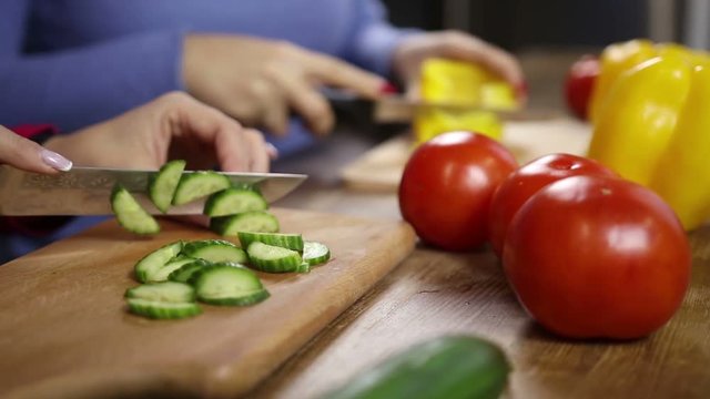 Two healthy middle-aged women cooking together in the kitchen. Closeup. Females hands slicing vegetables for fresh salad on wooden cutting boards. Home cooking and healthy eating. Slo mo. Dolly.