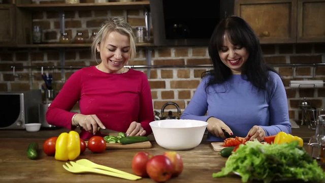 Two charming middle-aged women preparing healthy salad together in the kitchen and chatting. Attractive female adding chopped cucumber into bowl while her sister slicing tomato on cutting board.