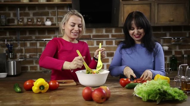 Joyful middle-aged woman stirring bowl of fresh salad while cooking healthy food together with her sister in modern kitchen. Healthy smiling women preparing lunch in the kitchen at home. Slo mo. Dolly