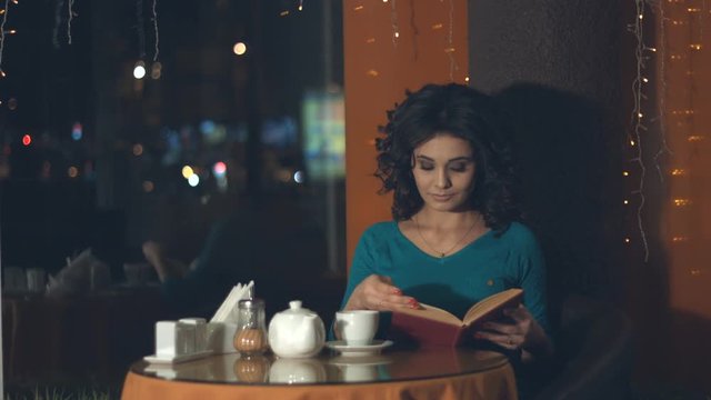Girl with a book in a cafe.
Slow motion. A pretty girl is sitting in a cafe and reading a book.
Outside the window lights the evening city.
