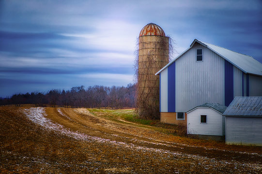 A quiet midwestern farmstead in winter, with glowing sky, silo and blue and white barn