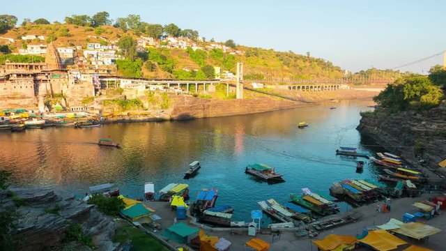 Sunset time lapse Omkareshwar cityscape, India, sacred hindu temple. Holy Narmada River, boats floating. Travel destination for tourists and pilgrims.
