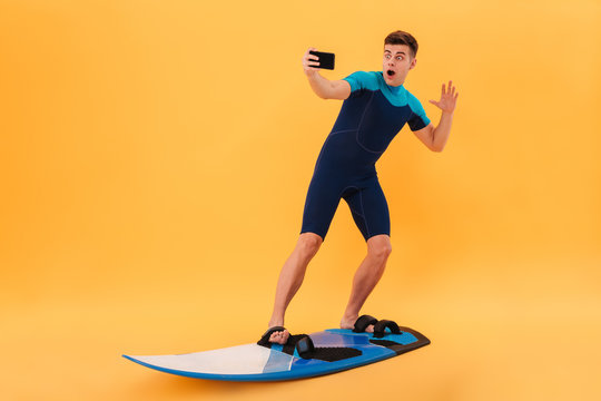 Full length photo of amased surfer guy taking selfie on smartphone while surfing on surfboard